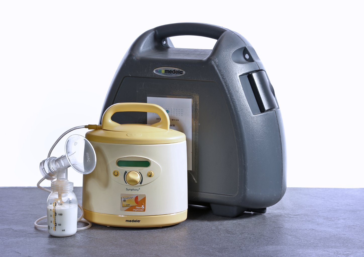 Eine Medela-Milchpumpe mit gelb-wei&szlig;em Motor, angeschlossenem Schlauch und Flasche steht neben einer gro&szlig;en, dunkelgrauen Tragetasche auf einer grauen Fl&auml;che.