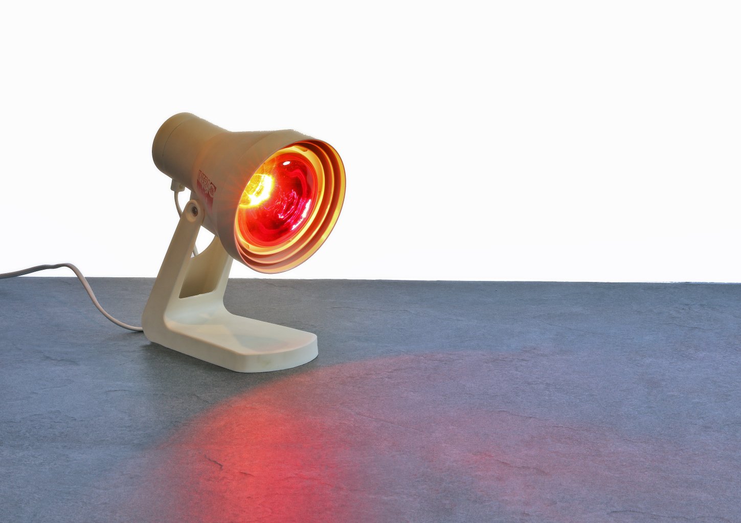 Eine kleine wei&szlig;e Infrarot-W&auml;rmelampe ist eingeschaltet und nach unten gerichtet und wirft ein warmes rotes Licht auf eine blaugraue Fl&auml;che. Der Hintergrund ist einfarbig wei&szlig;.