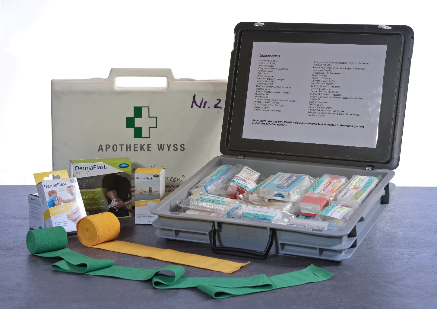 Auf einem Tisch steht ein Erste-Hilfe-Kasten mit verschiedenen medizinischen Hilfsmitteln, darunter Pflaster, Wickel und Kartons mit Pflastern. Der Kasten ist offen, mit ordentlich geordneten Gegenst&auml;nden im Inneren und einer Checkliste auf dem Deckel.