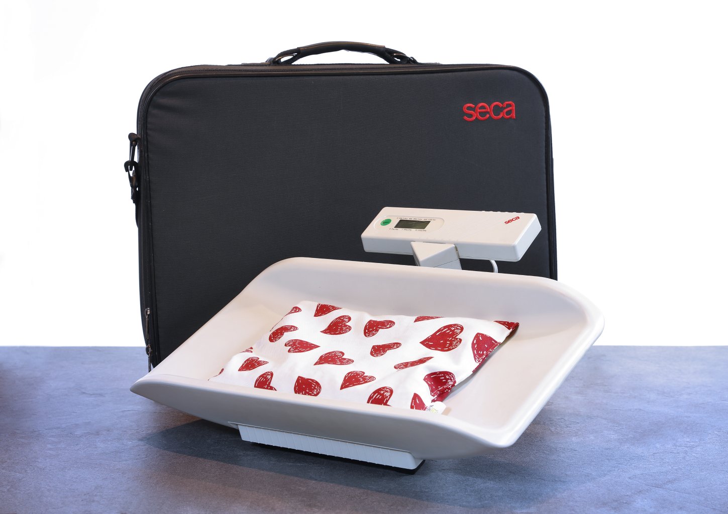 Eine tragbare S&auml;uglingswaage mit digitaler Anzeige, deren Schale mit einem wei&szlig;en, mit roten Herzen verzierten Tuch ausgekleidet ist, steht auf einem Tisch vor einer schwarzen Tragetasche mit der Aufschrift seca.