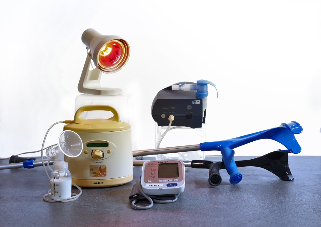 Eine Gruppe medizinischer Ger&auml;te auf einer grauen Fl&auml;che, darunter eine Milchpumpe, ein Blutdruckmessger&auml;t, eine blaue Greifzange, eine Lampe mit rotem Licht und ein Beatmungsger&auml;t, vor einem wei&szlig;en Hintergrund.