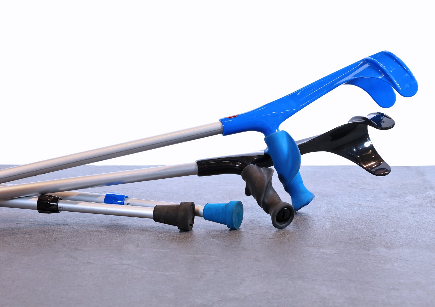 Zwei Unterarmgehst&uuml;tzen mit blauen Griffen und Armst&uuml;tzen liegen auf einer grauen Fl&auml;che. Die Kr&uuml;cken sind nebeneinander angeordnet, wobei ihre Spitzen nach links zeigen. Der Hintergrund ist wei&szlig;.