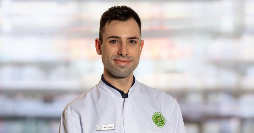 Ein Mann mit kurzen dunklen Haaren, der eine wei&szlig;e Kochjacke mit einem gr&uuml;nen kreisf&ouml;rmigen Logo und einem Namensschild mit der Aufschrift "Reto Oeschger" tr&auml;gt, steht vor einem verschwommenen Innenhintergrund und l&auml;chelt leicht in die Kamera.