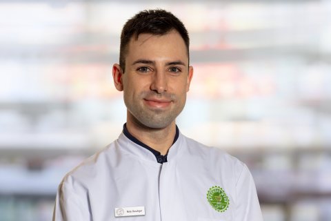 Ein Mann mit kurzen dunklen Haaren, der eine wei&szlig;e Kochjacke mit einem gr&uuml;nen kreisf&ouml;rmigen Logo und einem Namensschild mit der Aufschrift "Reto Oeschger" tr&auml;gt, steht vor einem verschwommenen Innenhintergrund und l&auml;chelt leicht in die Kamera.