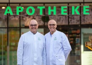 Zwei l&auml;chelnde M&auml;nner in wei&szlig;en Laborkitteln stehen vor einer Apotheke, &uuml;ber der ein gro&szlig;es gr&uuml;nes APOTHEKE-Schild zu sehen ist.