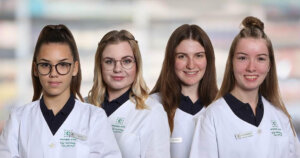 Vier junge Frauen in wei&szlig;en Laborkitteln und dunklen Hemden stehen nebeneinander und l&auml;cheln in die Kamera. Jede von ihnen tr&auml;gt ein Namensschild und hat verschiedene Frisuren. Hinter ihnen ist ein unscharfer Innenhintergrund zu sehen.