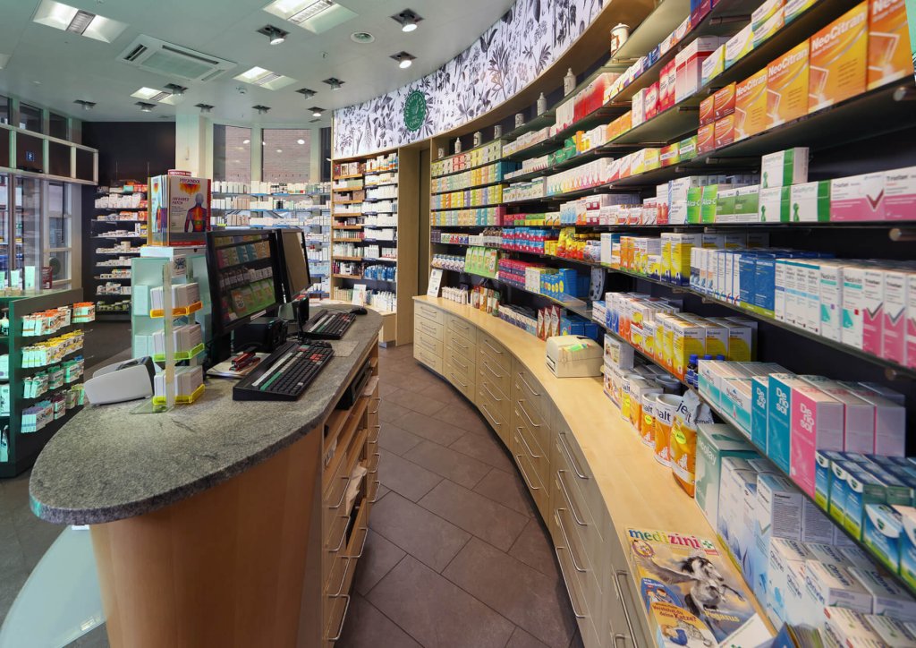 Eine moderne Apotheke mit geschwungenen Regalen, die mit verschiedenen Medikamenten und Gesundheitsprodukten best&uuml;ckt sind, einer Theke mit Computerbildschirmen und gro&szlig;en Fenstern, durch die Tageslicht f&auml;llt.