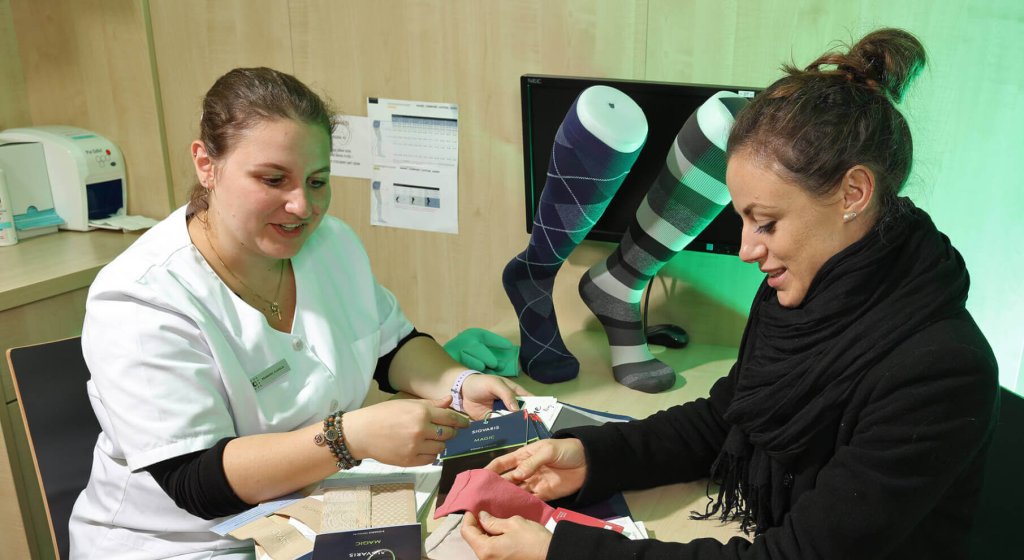 Eine medizinische Fachkraft zeigt einer Frau an einem Schreibtisch Stoffmuster, w&auml;hrend im Hintergrund zwei Schaufensterpuppen Beine mit gemusterten Kompressionsstr&uuml;mpfen zeigen. Beide Frauen l&auml;cheln und diskutieren &uuml;ber die besten Optionen zur Anpassung von Kompressionsstr&uuml;mpfen.