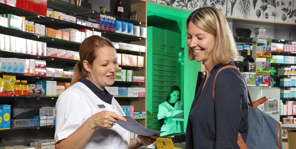Ein Apotheker zeigt einem l&auml;chelnden Reisenden in einer Apotheke ein Dokument. Im Hintergrund stehen Regale mit verschiedenen Medikamenten, und in einem Raum dahinter ist ein weiterer Mitarbeiter zu sehen.