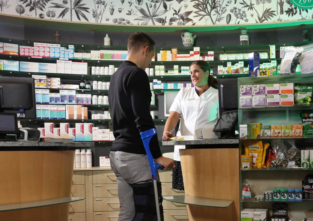 Ein Mann mit Kr&uuml;cken steht an einem Apothekentresen und spricht mit einem l&auml;chelnden Apotheker in einem wei&szlig;en Kittel. Die Regale hinter dem Tresen sind mit Medikamenten, Gesundheitsprodukten und Sanit&auml;tsartikeln gef&uuml;llt.