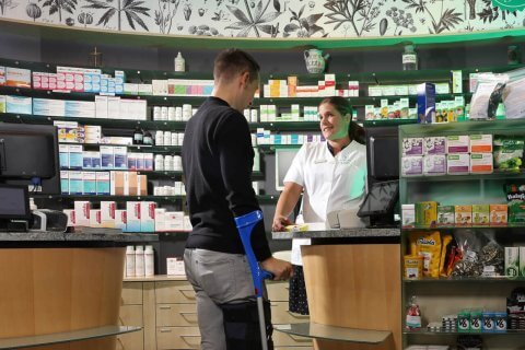 Ein Mann mit Kr&uuml;cken steht an einem Apothekentresen und spricht mit einem l&auml;chelnden Apotheker in einem wei&szlig;en Kittel. Die Regale hinter dem Tresen sind mit Medikamenten, Gesundheitsprodukten und Sanit&auml;tsartikeln gef&uuml;llt.