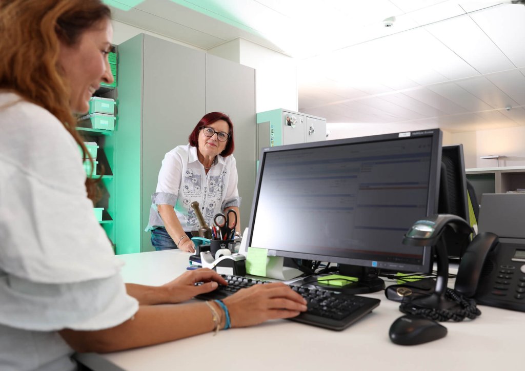 Zwei Frauen arbeiten in einem hellen B&uuml;ro; die eine tippt an einem Computer, w&auml;hrend die andere neben dem Schreibtisch steht und sie anschaut. Auf dem Schreibtisch im Vordergrund sind B&uuml;romaterial und ein Monitor zu sehen.