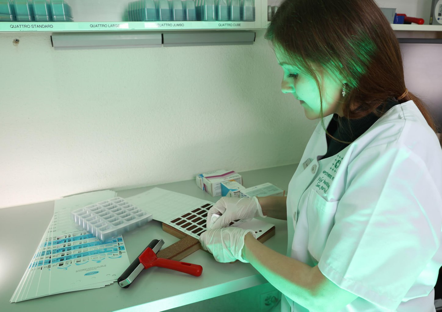 Eine Frau in Laborkittel und Handschuhen untersucht Proben an einem Laborarbeitsplatz, benutzt Werkzeuge und dokumentiert die Ergebnisse. Auf dem Schreibtisch sind Tabletts, ein Hammer, Formulare und ein Medikamenten-Dosiersystem unter grün getöntem Licht zu sehen.