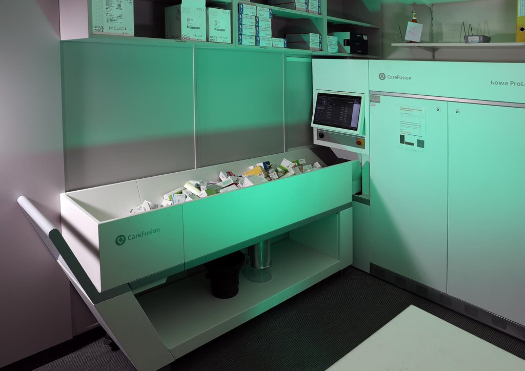 Ein Medikamentenausgabeautomat mit Touchscreen und offenem Tablett mit verschiedenen Medikamenten steht in einem Klinikraum am Standort Baden, dar&uuml;ber Regale mit Boxen, darunter Schr&auml;nke, beleuchtet mit gr&uuml;nem Licht.