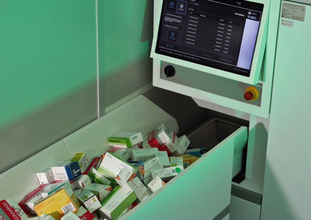 Ein Apothekenautomat am Standort Baden zeigt einen digitalen Bildschirm an und gibt zahlreiche Medikamentenschachteln in einen darunter liegenden Beh&auml;lter aus, um ein organisiertes und effizientes Medikamentenmanagement zu demonstrieren.