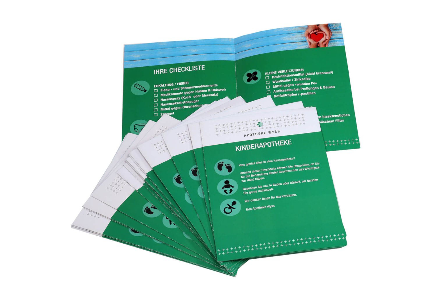 Ein Satz gr&uuml;ner Informationsbrosch&uuml;ren in deutscher Sprache mit dem Titel "KINDERAPOTHEKE", aufgef&auml;chert mit Checklisten, Ferien-Check-Symbolen und einem aufgeschlagenen Heft mit einer Checkliste und einigen Bildern.