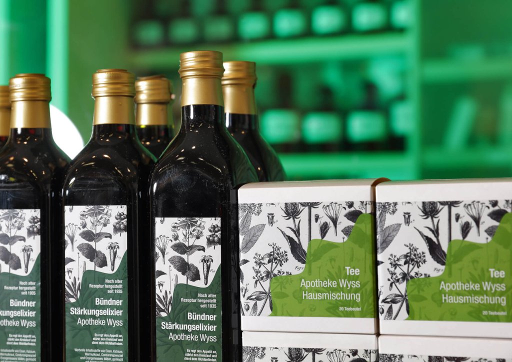 Flaschen und Schachteln mit gr&uuml;nen und wei&szlig;en Kr&auml;uteretiketten stehen auf einer Theke in einer Apotheke oder einem Kr&auml;uterladen, wobei die Regale mit den Tiegeln und Produkten im gr&uuml;n beleuchteten Hintergrund verschwimmen.