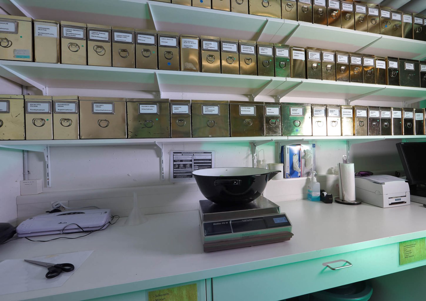 Ein Laborarbeitsplatz mit einer Digitalwaage, einer schwarz-wei&szlig;en Sch&uuml;ssel, Scheren, Papieren und verschiedenen Ger&auml;ten auf dem Tresen. Dar&uuml;ber befinden sich Regale mit vielen beschrifteten Metallbeh&auml;ltern mit runden Griffen.
