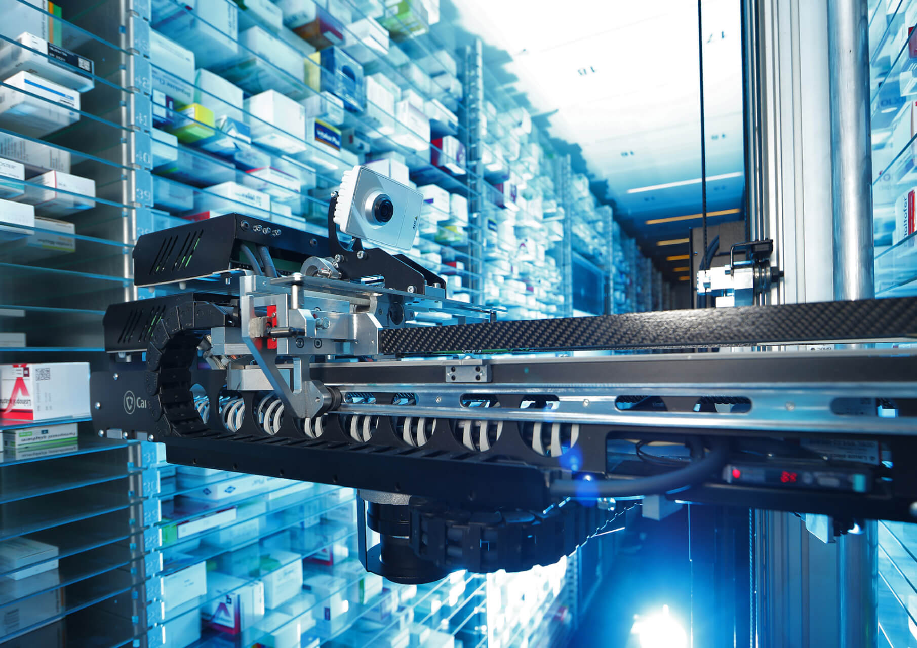 Ein Roboterarm ordnet Medikamentenboxen in einem modernen, automatisierten Apothekenlager am Standort Baden. Im Hintergrund stehen Regale mit verschiedenen Medikamentenpackungen und eine helle, fokussierte Beleuchtung.