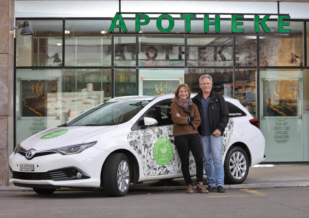 Eine l&auml;chelnde Frau und ein l&auml;chelnder Mann stehen vor einem wei&szlig;en Auto mit gr&uuml;ner Aufschrift, das f&uuml;r ihren Hauslieferservice wirbt und vor einem Geb&auml;ude mit einem gro&szlig;en APOTHEKE-Schild &uuml;ber dem Eingang geparkt ist.