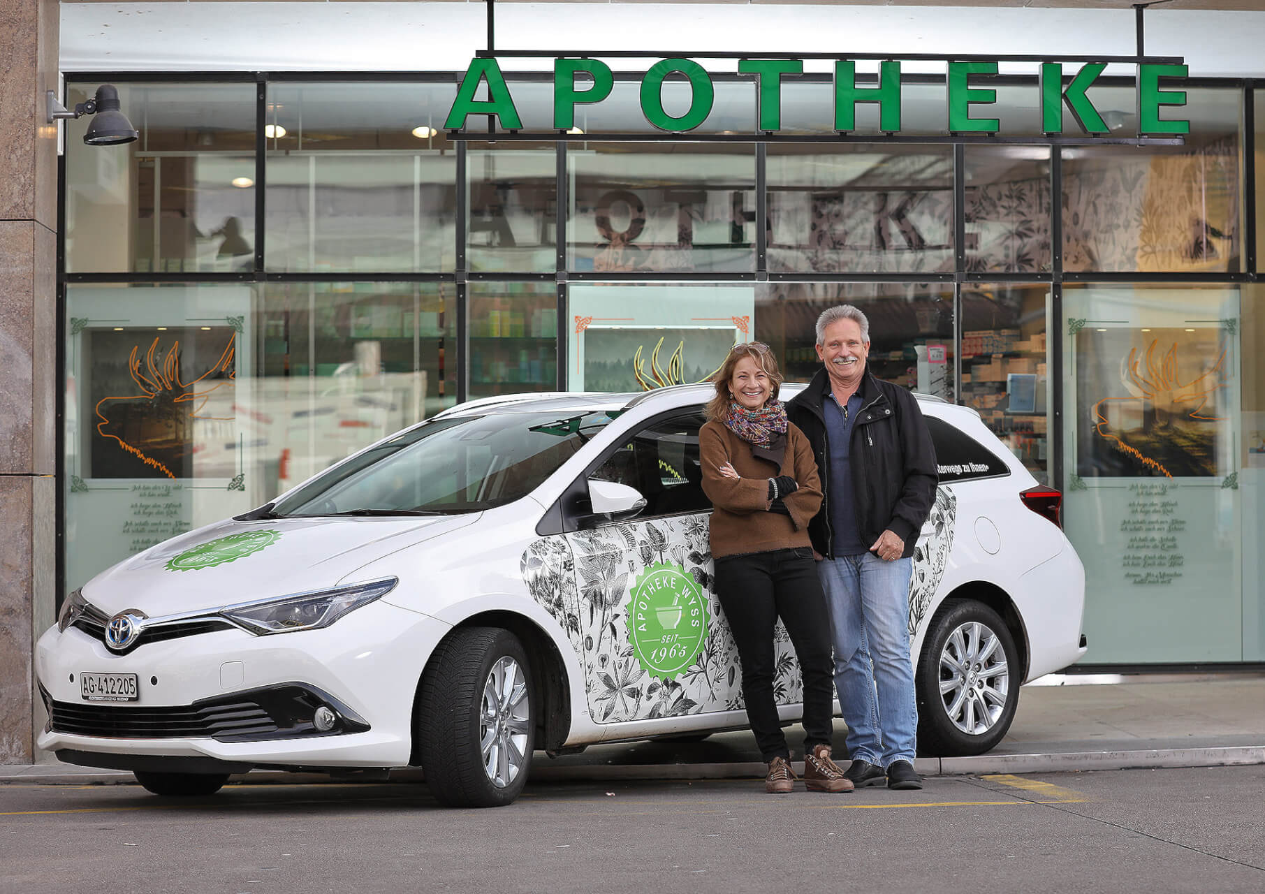 Eine l&auml;chelnde Frau und ein l&auml;chelnder Mann stehen vor einem wei&szlig;en Auto mit gr&uuml;ner Aufschrift, das f&uuml;r ihren Hauslieferservice wirbt und vor einem Geb&auml;ude mit einem gro&szlig;en APOTHEKE-Schild &uuml;ber dem Eingang geparkt ist.