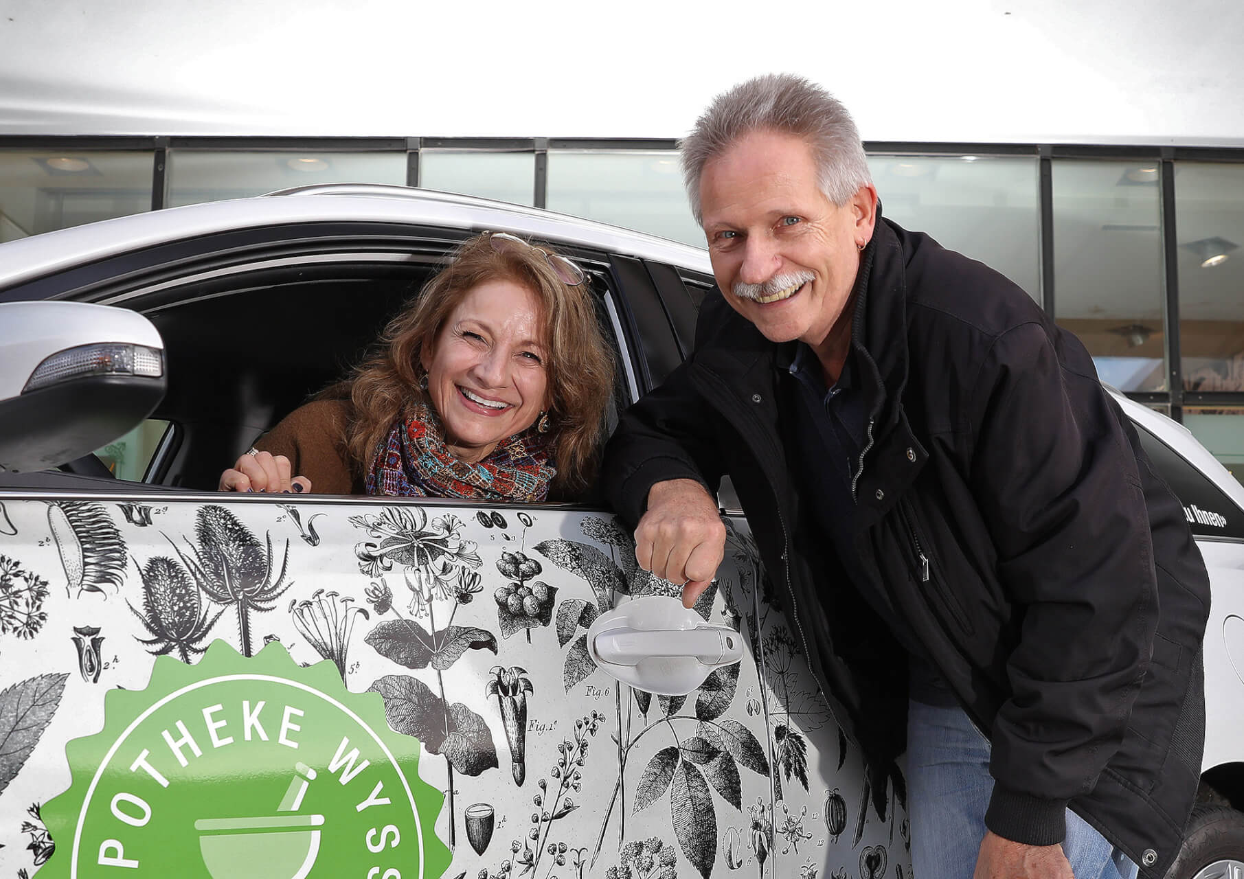 Eine l&auml;chelnde Frau sitzt in einem mit botanischen Illustrationen geschm&uuml;ckten Auto, das einen Hauslieferservice darstellt, w&auml;hrend ein freundlicher Mann neben ihr steht und den T&uuml;rgriff h&auml;lt. Beide wirken fr&ouml;hlich und sympathisch.