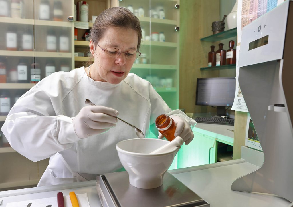 Eine Frau in einem Laborkittel und mit Handschuhen sch&uuml;ttet Pulver aus einer braunen Flasche der Wyss Manufaktur in einen M&ouml;rser und h&auml;lt dabei einen Metallspatel in der Hand. Sie befindet sich in einer Apotheke oder einem Labor mit Regalen voller Flaschen und einem Computer im Hintergrund.