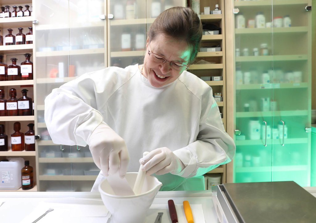Eine Person in wei&szlig;em Laborkittel und mit Handschuhen l&auml;chelt, w&auml;hrend sie in einer Apotheke oder einem Labor der Wyss Manufaktur einen M&ouml;rser und einen St&ouml;&szlig;el benutzt, w&auml;hrend im Hintergrund Regale mit Flaschen und Beh&auml;ltern zu sehen sind.