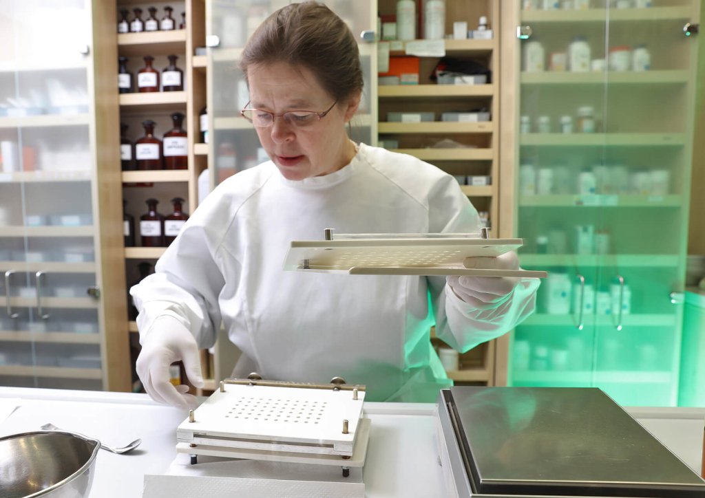 Eine Frau in Laborkittel und Handschuhen montiert in einem Labor eine Abf&uuml;llmaschine f&uuml;r Tablettenkapseln. Im Hintergrund sind Regale mit Flaschen und Beh&auml;ltern zu sehen, die Schweizer Handwerkskunst der Wyss Manufaktur demonstrieren.