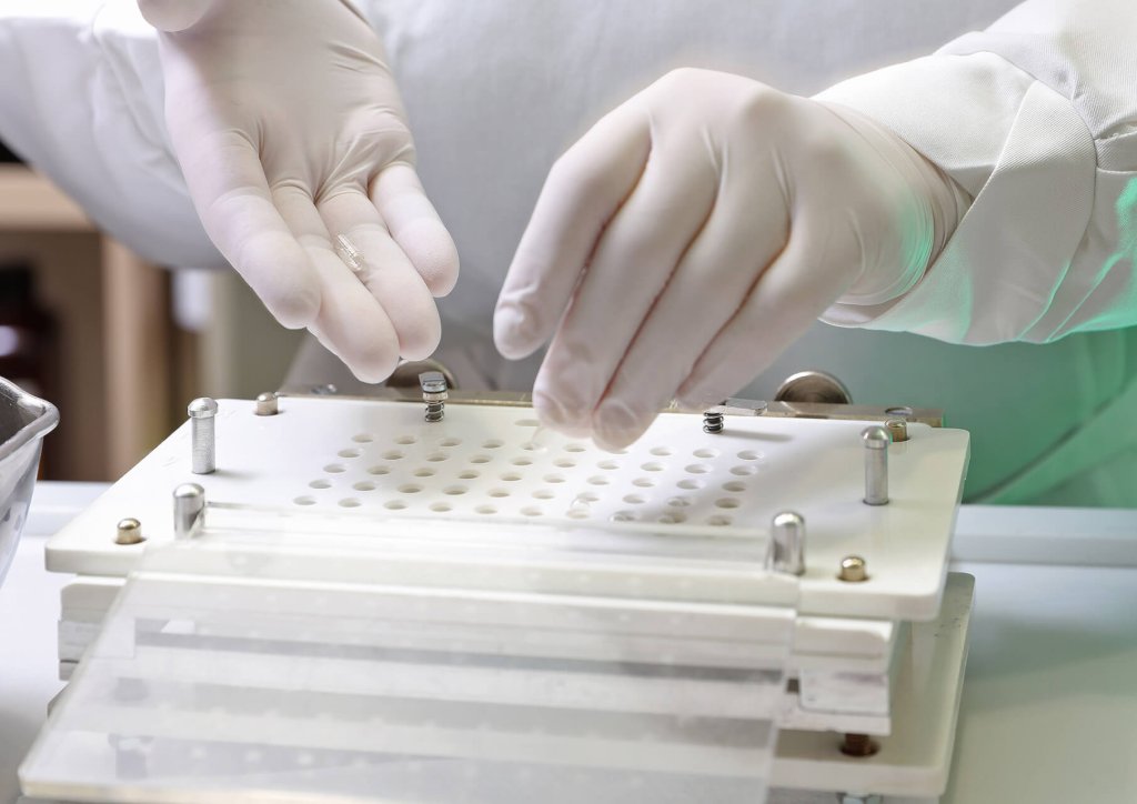 Eine Person mit weissen Handschuhen f&uuml;llt in einem sauberen Labor oder in einer Apotheke Kapseln mit einer Pillenmaschine der Wyss Manufaktur mit mehreren L&ouml;chern.