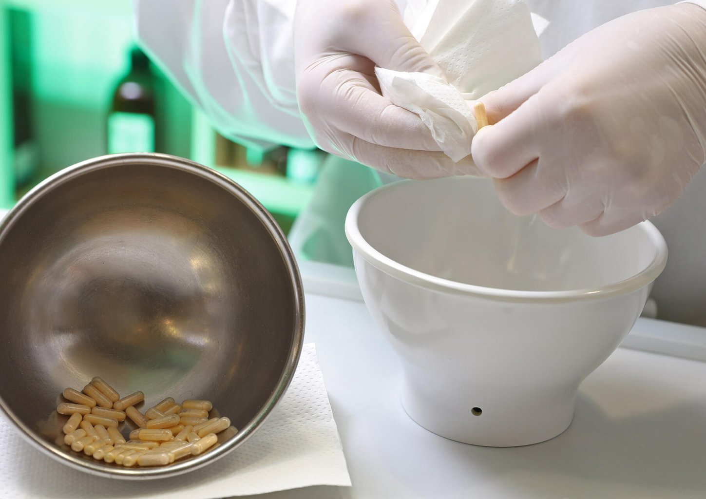 Eine Person mit wei&szlig;en Handschuhen bereitet an einem Labortisch der Wyss Manufaktur Kapseln vor. Sie h&auml;lt ein Taschentuch in der Hand und arbeitet mit einem wei&szlig;en M&ouml;rser und St&ouml;&szlig;el neben einer Metallsch&uuml;ssel mit gelblichen Kapseln.