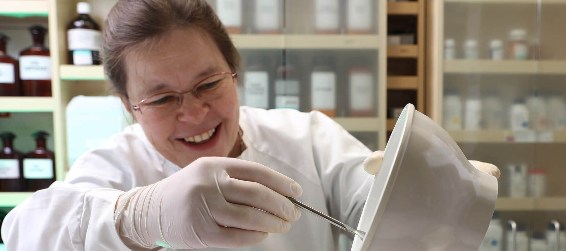 Eine l&auml;chelnde Person im Laborkittel und mit Handschuhen mischt in der Wyss Manufaktur Substanzen in einer wei&szlig;en Sch&uuml;ssel mit einem Spatel, umgeben von Regalen mit Flaschen und Gl&auml;sern in einer Laborumgebung.