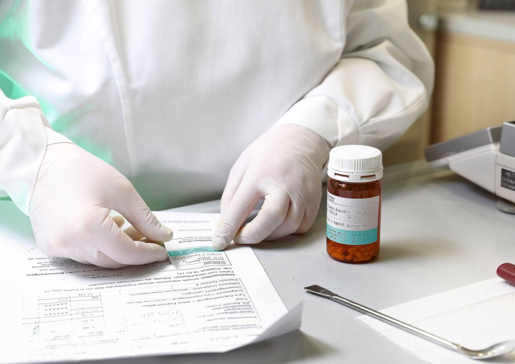 Eine Person in einem wei&szlig;en Laborkittel und mit Handschuhen pr&uuml;ft Papierkram an einem Schreibtisch mit einer verschreibungspflichtigen Tablettenflasche der Wyss Manufaktur, einem Metallwerkzeug und einer Waage, was auf eine medizinische oder Laborumgebung schlie&szlig;en l&auml;sst.