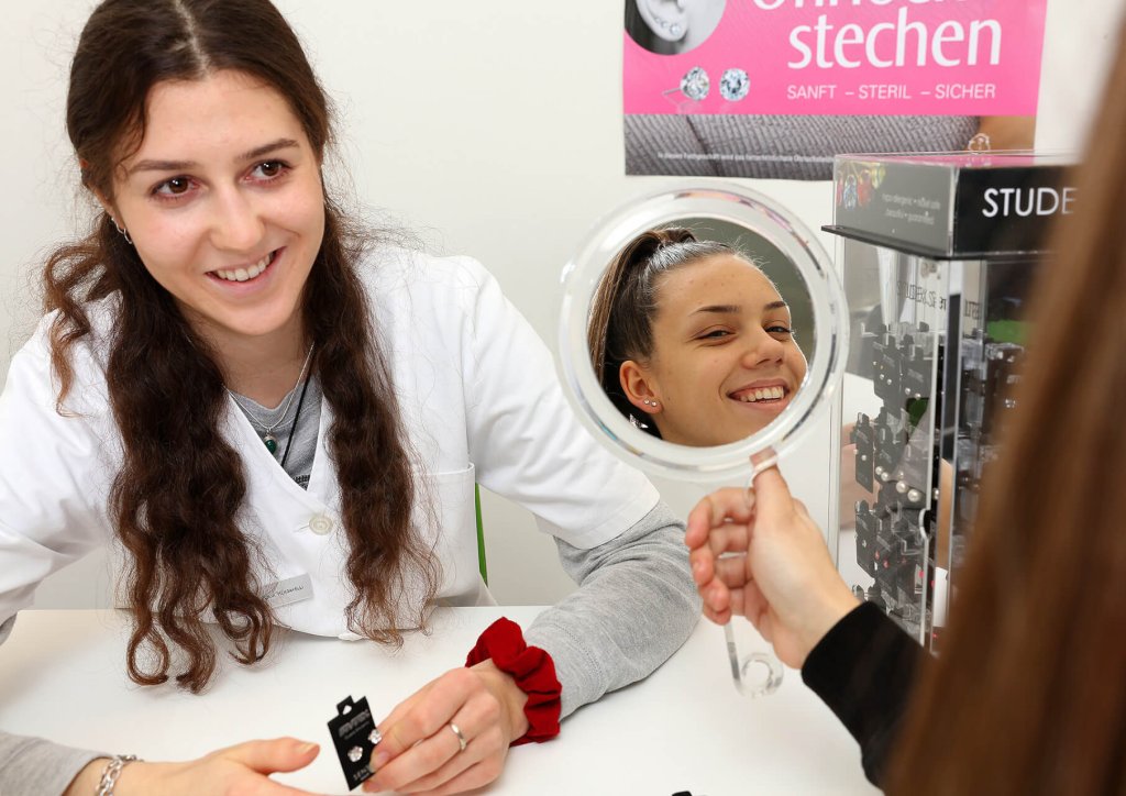 Eine Frau in einem wei&szlig;en Kittel l&auml;chelt, w&auml;hrend eine andere Frau einen runden Spiegel h&auml;lt, in dem sich ihr gl&uuml;ckliches Gesicht spiegelt. Sie scheinen in einer Klinik oder einem Juweliergesch&auml;ft zu sein, wo sie m&ouml;glicherweise &uuml;ber Ohrlochstechen sprechen.