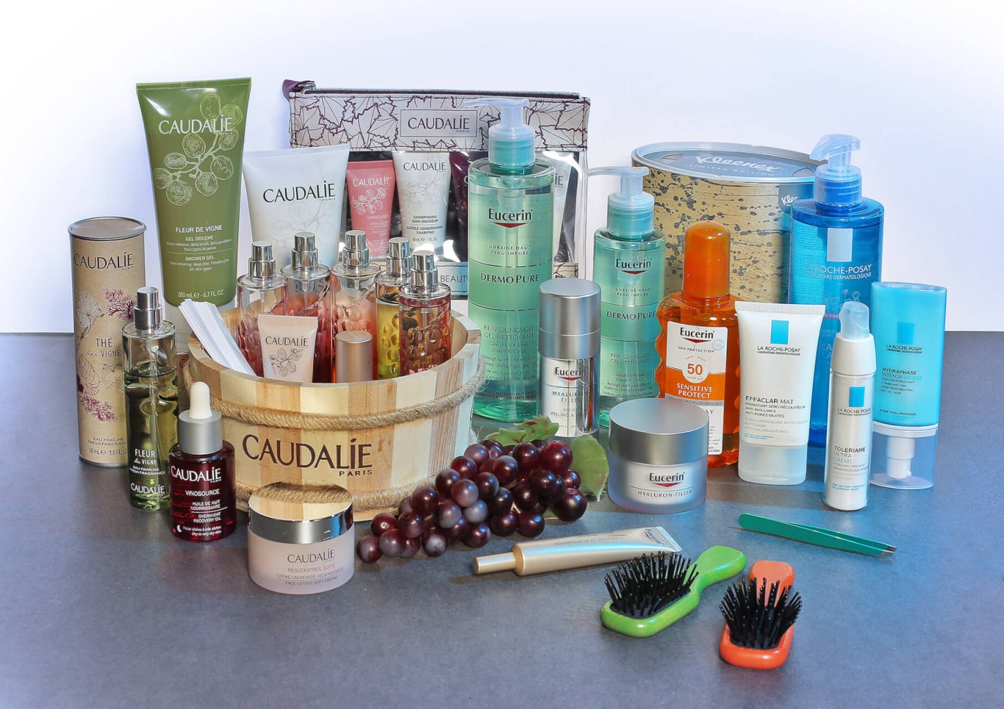 Verschiedene Hautpflegeprodukte von Caudalie, Eucerin und La Roche-Posay, Parfums, Cremes und Lotionen sind auf einer grauen Fl&auml;che mit zwei Haarb&uuml;rsten, einer Weintraube und einem Holzkorb angeordnet.