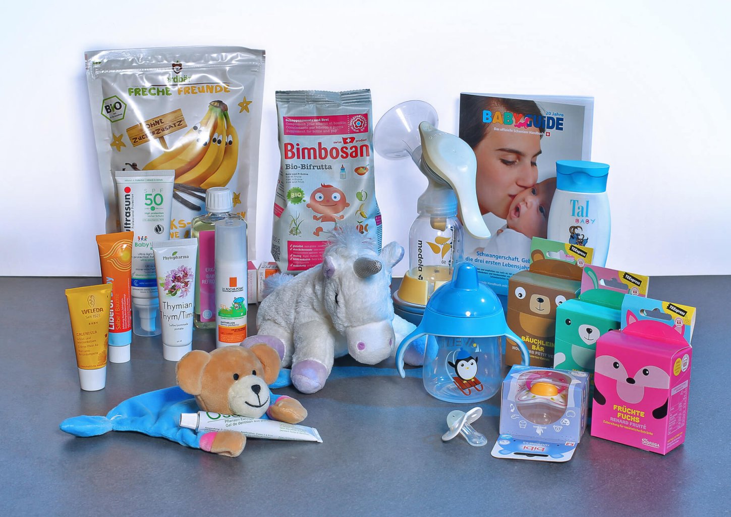 Auf einer grauen Fl&auml;che sind verschiedene Babypflegeprodukte, Spielzeuge und Lebensmittel angeordnet, darunter Flaschen, Cremes, Schnuller, ein Pl&uuml;schb&auml;r, ein Pl&uuml;scheinhorn und bunte Verpackungen mit Tiermotiven.