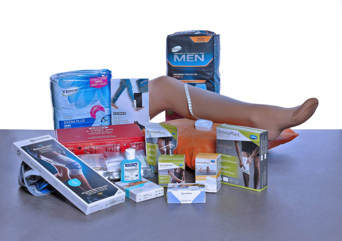 Eine Vielzahl von Sanit&auml;tsartikeln und K&ouml;rperpflegeprodukten, darunter Windeln f&uuml;r Erwachsene, Kompressionsstr&uuml;mpfe (ausgestellt an einem Bein einer Schaufensterpuppe), Wundauflagen, eine Kniest&uuml;tze, Einweghandschuhe und antiseptische L&ouml;sung, angeordnet auf einer grauen Fl&auml;che.