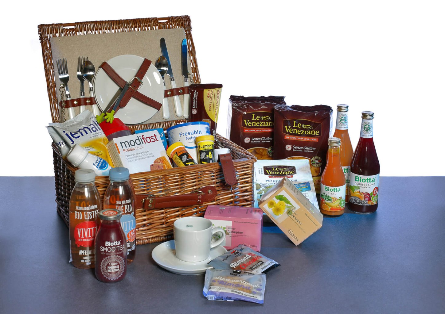 Ein Picknickkorb, gef&uuml;llt mit Tellern, Besteck und verschiedenen Lebensmitteln, steht offen auf einem Tisch, umgeben von Getr&auml;nken, verpackten Snacks, P&uuml;lverchen und einer wei&szlig;en Tasse mit Untertasse. Die Flaschen und P&auml;ckchen sind fein s&auml;uberlich angeordnet.