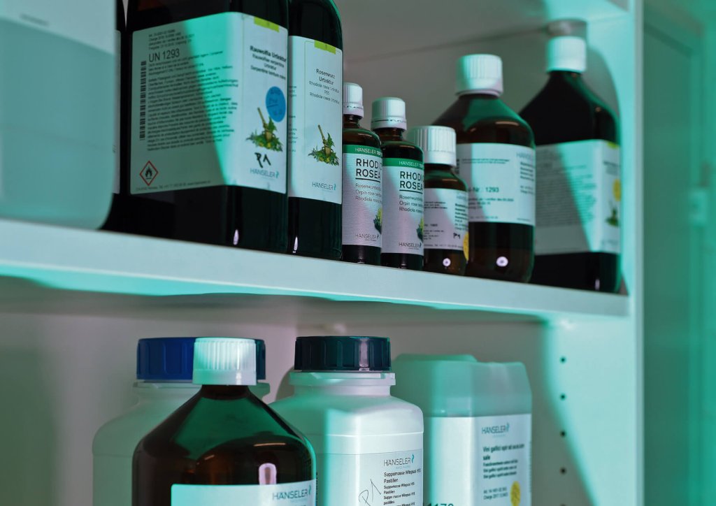 Verschiedene Flaschen und Beh&auml;lter mit Chemikalien und L&ouml;sungen sind auf wei&szlig;en Regalen angeordnet, die jeweils mit Text und Grafiken beschriftet sind. Ein blaugr&uuml;nes Licht beleuchtet die Szene und wirft farbige Glanzlichter auf die Oberfl&auml;chen.