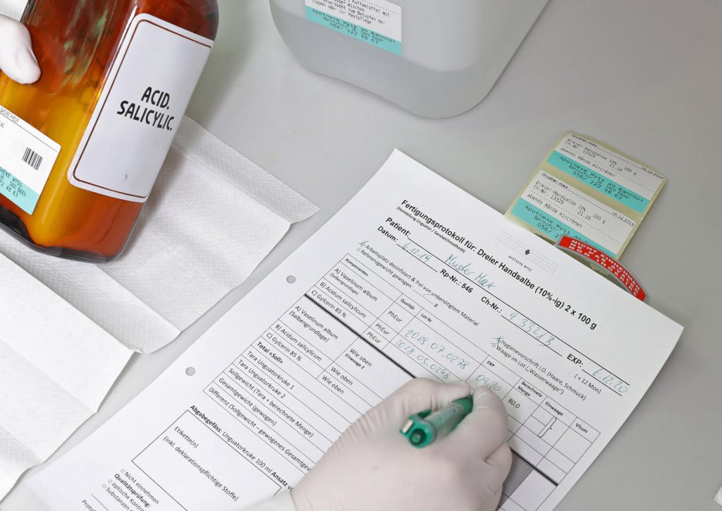 Eine behandschuhte Hand f&uuml;llt ein pharmazeutisches Formular auf einem Klemmbrett aus, das neben einer gro&szlig;en braunen Flasche der Wyss Manufaktur mit der Aufschrift Acid Salicylic und anderen Laborbeh&auml;ltern auf einer wei&szlig;en Fl&auml;che steht.