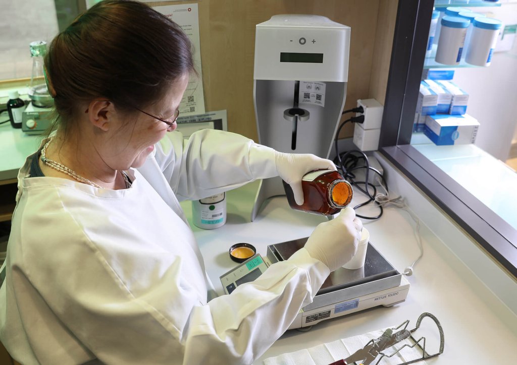 Eine Person im Laborkittel und mit Handschuhen gie&szlig;t Fl&uuml;ssigkeit aus einer braunen Flasche der Wyss Manufaktur auf eine digitale Waage in einem Labor, in dem Ger&auml;te und Zubeh&ouml;r auf dem Tresen stehen.
