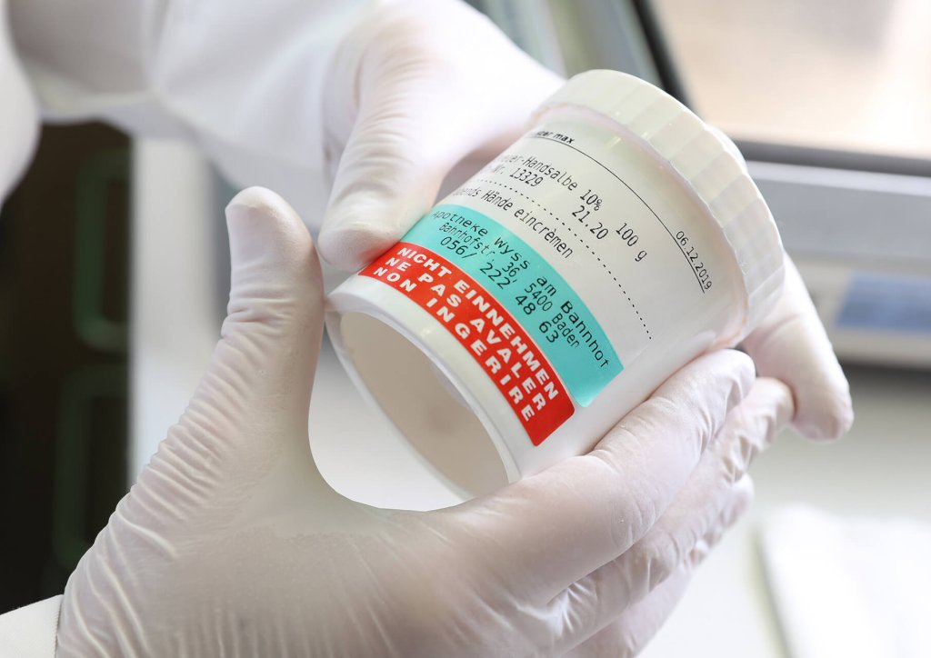 Eine Person mit wei&szlig;en Handschuhen h&auml;lt ein wei&szlig;es Beh&auml;ltnis der Wyss Manufaktur mit mehreren Etiketten und gedrucktem Text, darunter ein rotes Warnetikett in deutscher Sprache und eine Dosierungsanleitung, in der N&auml;he eines Fensters.