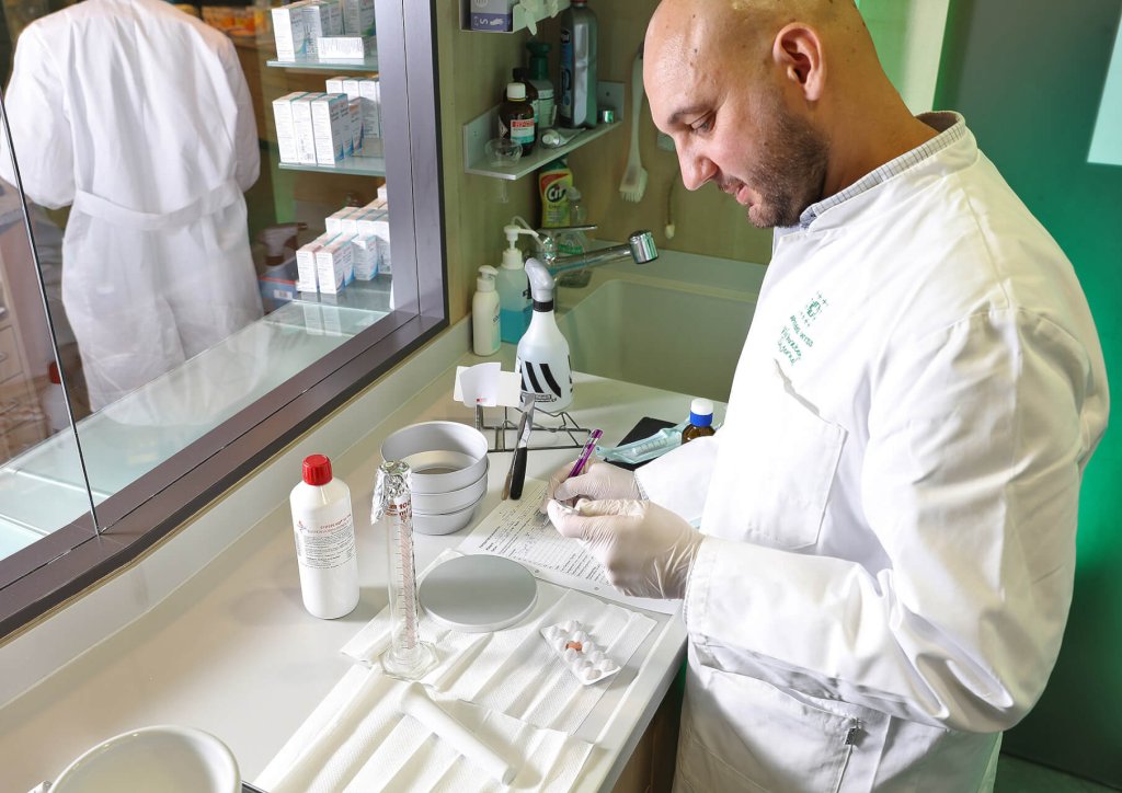 Ein Mann in wei&szlig;em Laborkittel und Handschuhen schreibt Notizen an einem Labortisch der Wyss Manufaktur mit wissenschaftlichen Ger&auml;ten, Glaswaren und Medikamentenflaschen; durch das Fenster ist eine weitere Person in einem Laborkittel zu sehen.