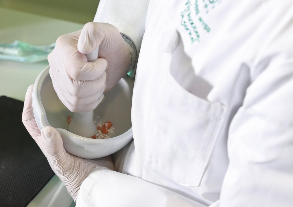 Eine Person mit wei&szlig;en Handschuhen und einem Laborkittel zerkleinert rote und wei&szlig;e Substanzen in einem wei&szlig;en M&ouml;rser mit einem St&ouml;&szlig;el, wahrscheinlich in einem Labor oder einer Apotheke der Wyss Manufaktur.