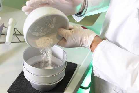 Eine Person, die einen Laborkittel und Handschuhe tr&auml;gt, sch&uuml;ttet ein wei&szlig;es Pulver aus einer Schale in einen Stapel von Sieben auf einem Labortisch der Wyss Manufaktur, wahrscheinlich zur Vorbereitung eines wissenschaftlichen Tests oder einer Qualit&auml;tskontrolle.