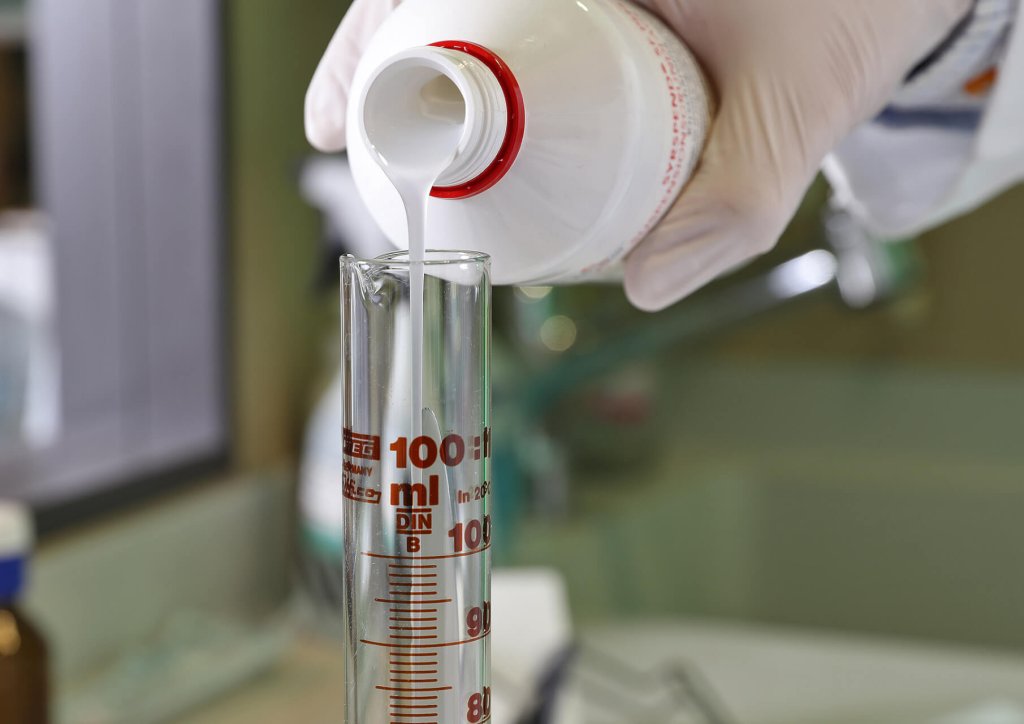 Eine behandschuhte Hand gie&szlig;t in einer Laborumgebung eine wei&szlig;e Fl&uuml;ssigkeit aus einer Flasche der Wyss Manufaktur in einen 100-ml-Messzylinder.