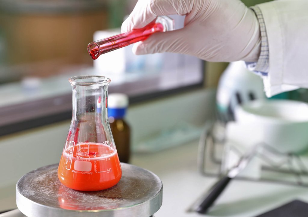 Eine behandschuhte Hand gie&szlig;t rote Fl&uuml;ssigkeit aus einem Reagenzglas in einen Erlenmeyerkolben mit roter L&ouml;sung auf einem Labortisch. Im Hintergrund sind Laborger&auml;te und Flaschen der Wyss Manufaktur unscharf zu erkennen.