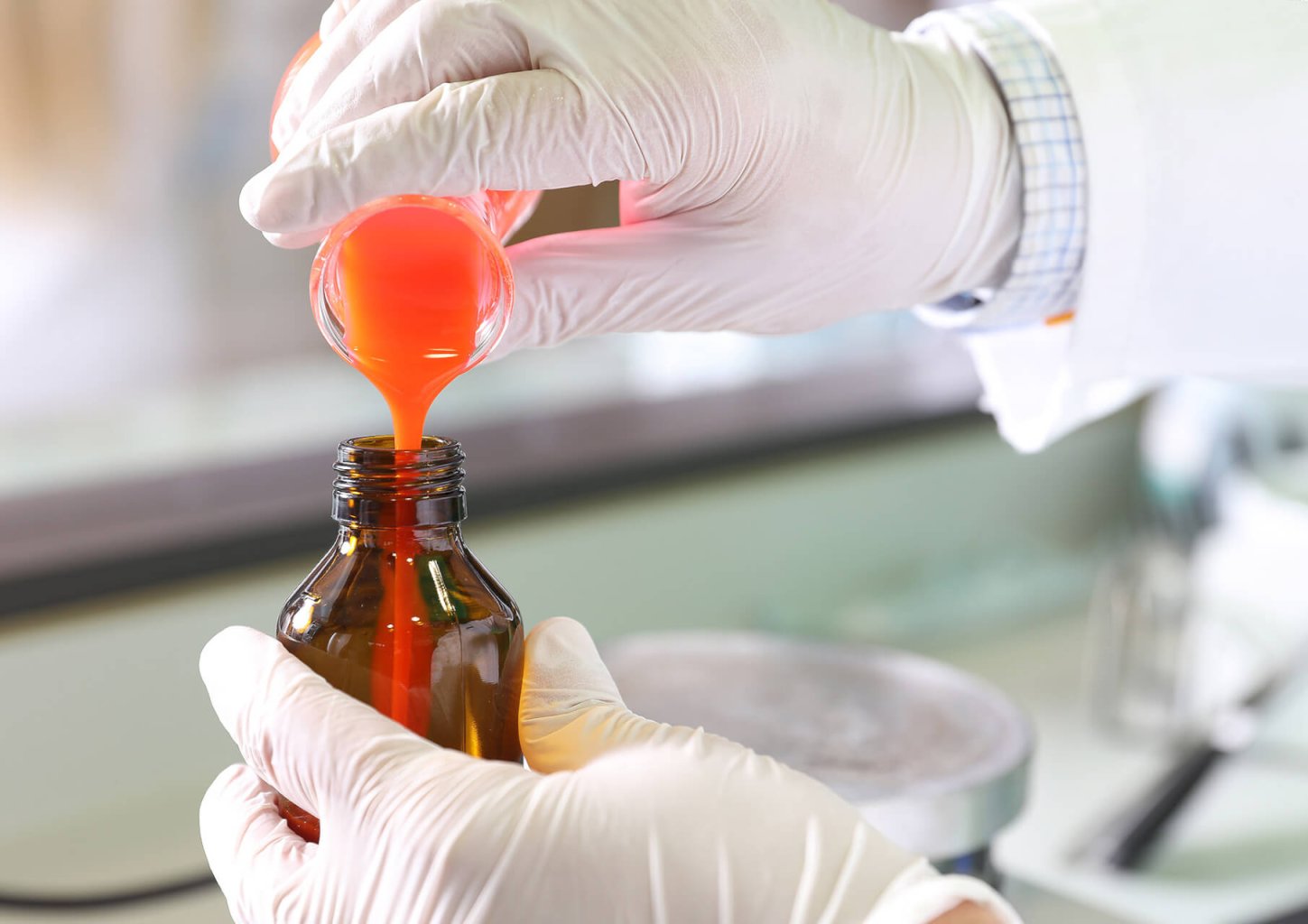 Eine Person mit weißen Handschuhen gießt eine orangefarbene Flüssigkeit aus einem Becherglas in eine braune Glasflasche, wahrscheinlich in einer Laborumgebung der Wyss Manufaktur.