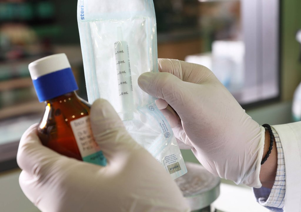 Eine Person mit wei&szlig;en Handschuhen h&auml;lt eine kleine braune Medikamentenflasche in der einen und eine unge&ouml;ffnete sterile Spritze in der anderen Hand in einer medizinischen oder Laborumgebung der Wyss Manufaktur.