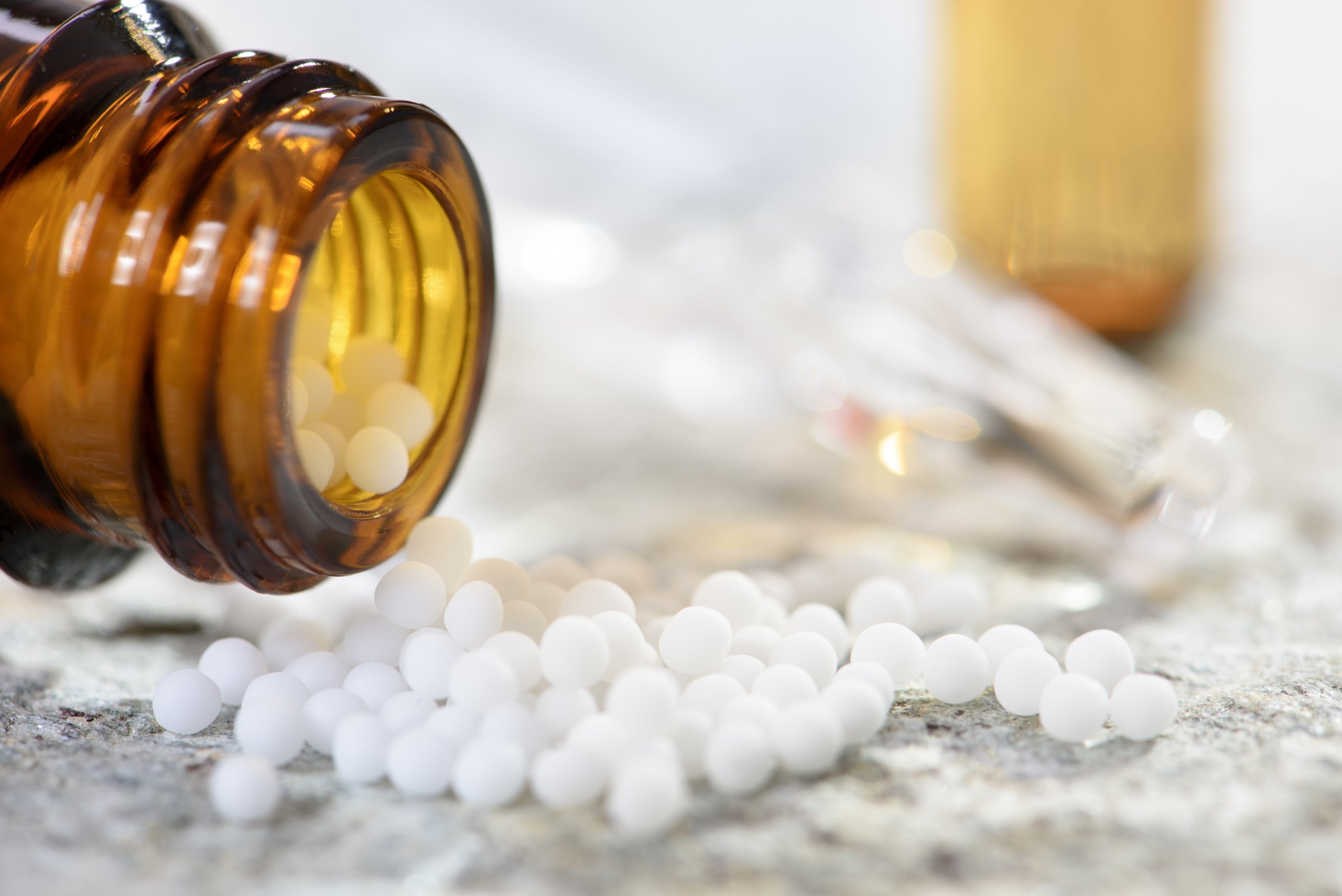 Eine braune Glasflasche ist umgekippt und hat kleine wei&szlig;e runde K&uuml;gelchen auf eine helle Fl&auml;che versch&uuml;ttet, mit verschwommenen Glasfl&auml;schchen im Hintergrund - eine h&auml;ufige Szene in der Praxis der Alternativmedizin.